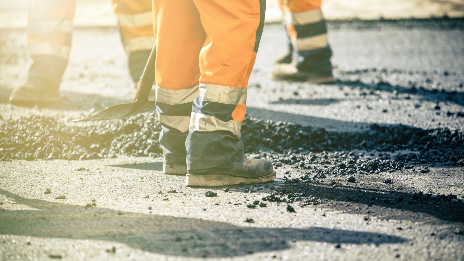 Workers are shown using shovels to fill pot holes on a roadway. They are wearing orange, reflective pants and workboots.