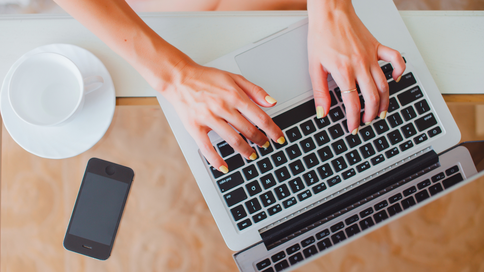 A woman types on a silver laptop surrounded by various pieces of technology including a smartphone. 