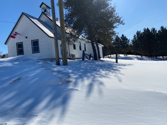 The photo shows a white building on a hill. This is the Snow Road Community Centre in North Frontenac. It is sunny outside and the ground is covered with snow. 