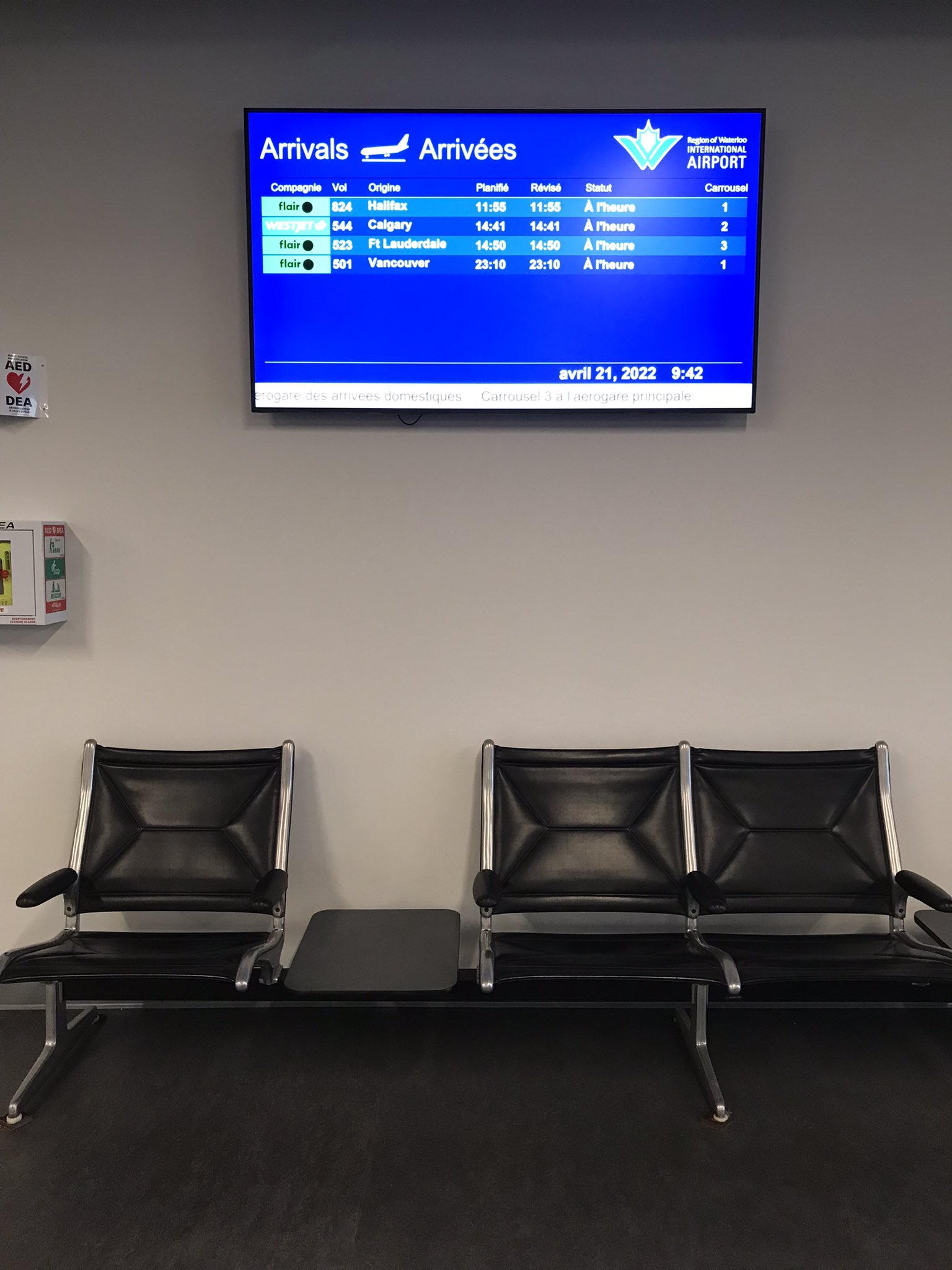 A photo of black chairs in the departure area of the Region of Waterloo International Airport, with a TV in the background showing the status of incoming and outgoing flights. 