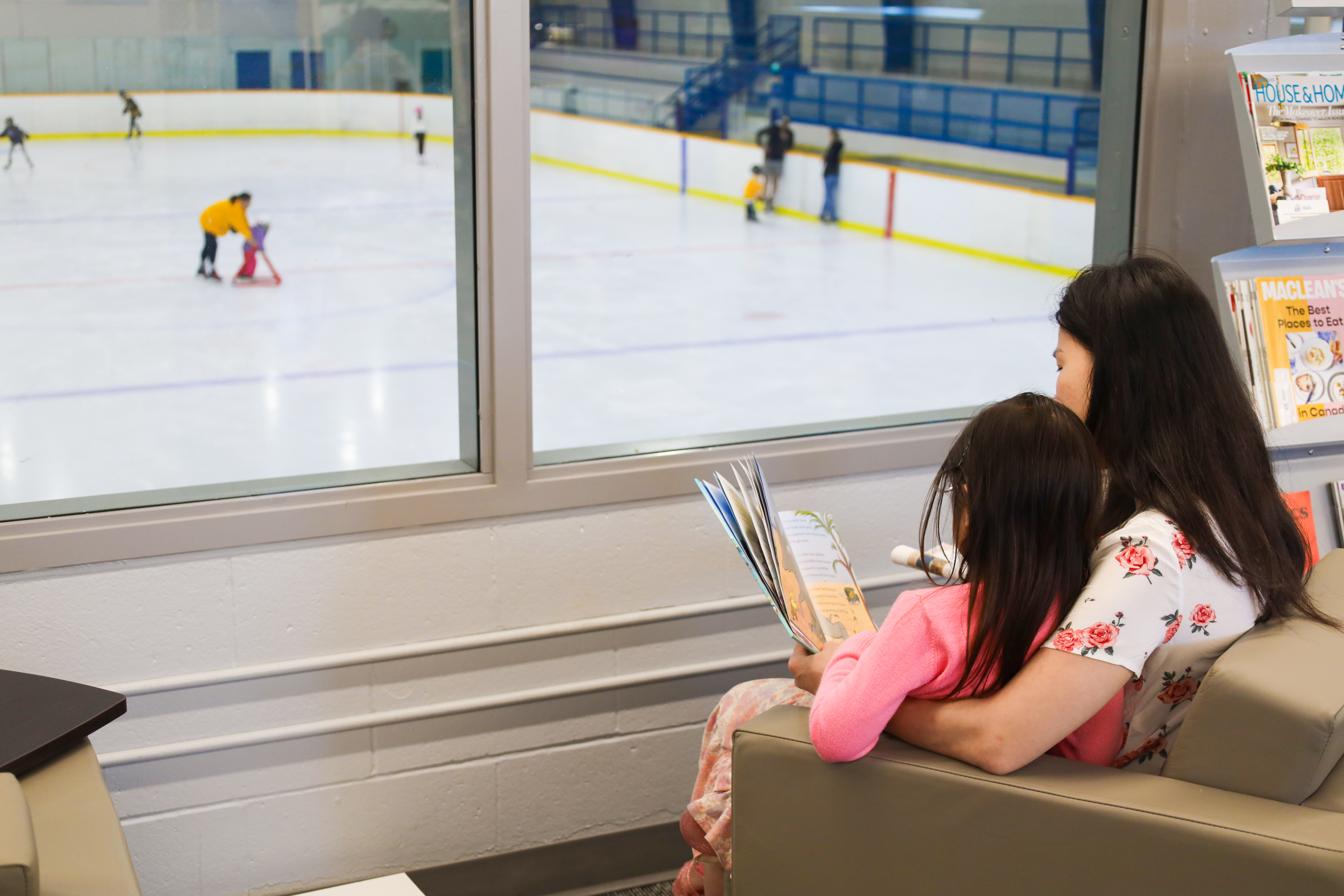 A parent and child reading a book while looking through a window at a skating rink below