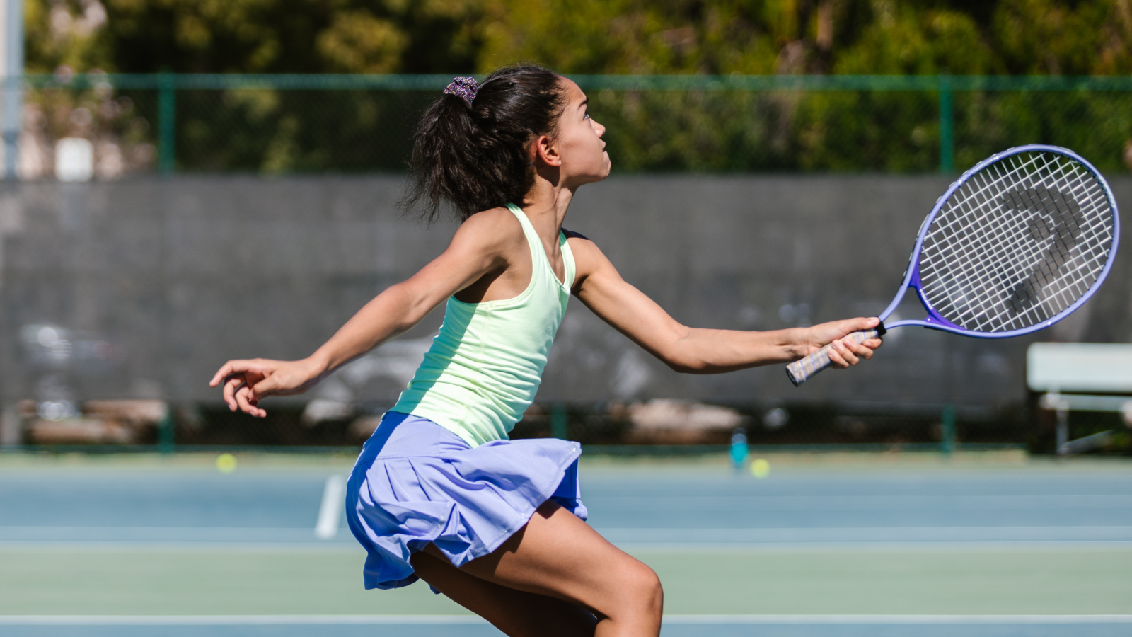 A woman playing tennis on a sunny day wearing a purple skirt and green tank top.