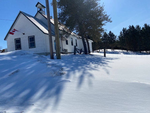 The photo shows a white building on a hill. This is the Snow Road Community Centre in North Frontenac. It is sunny outside and the ground is covered with snow.