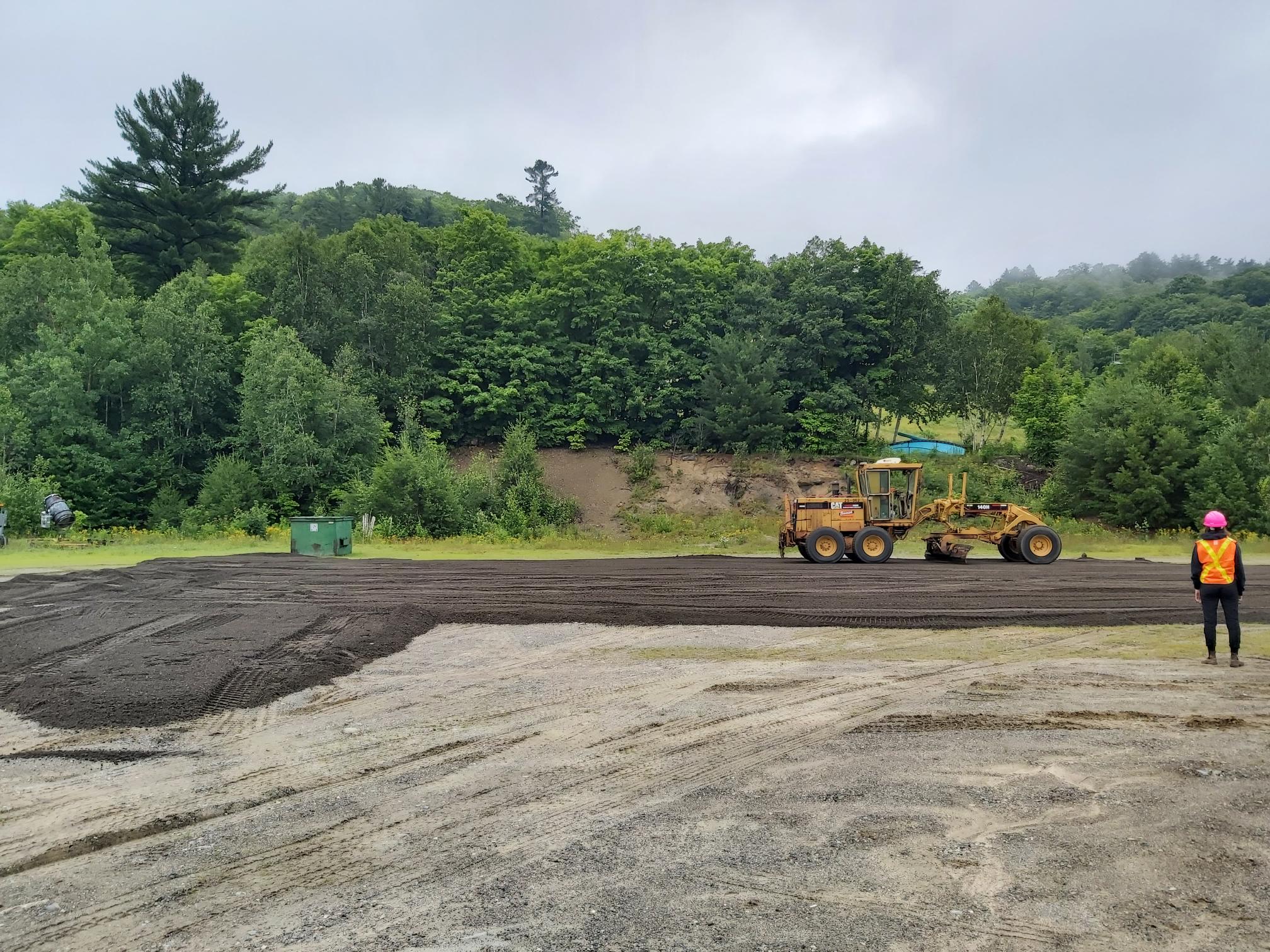 A construction worker watches as machinery smooths asphalt in a ski hill parking lot.