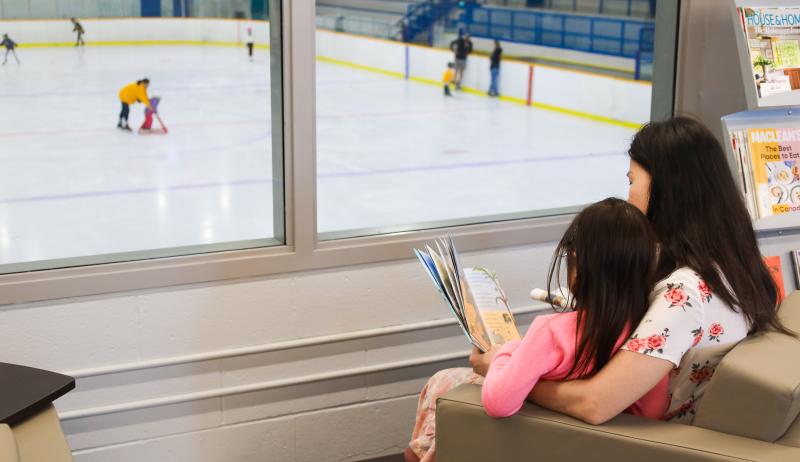 A parent and child reading a book beside a window to a skating rink