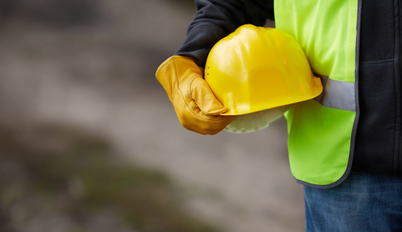 A construction worker stands holding a yellow helmet. 