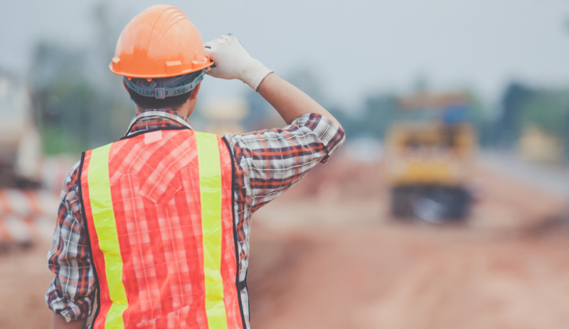 A construction worker wearing an orange helmet and vest surveys a job site. 
